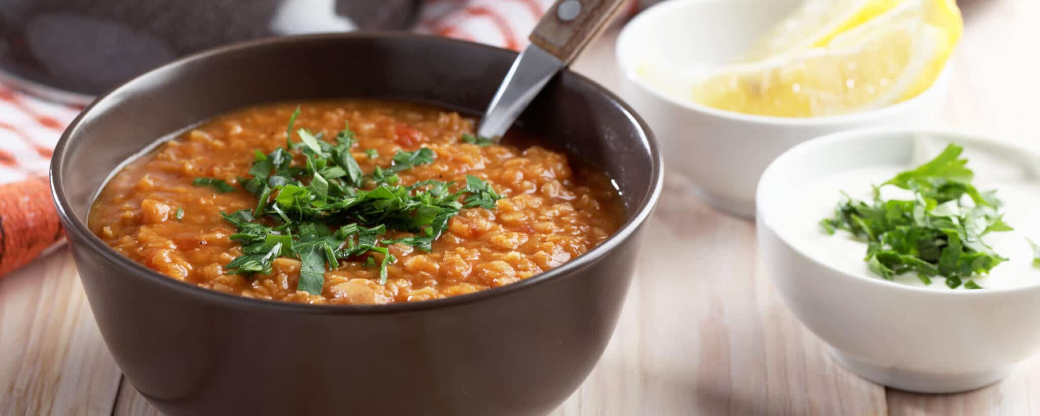 Lentil soup topped with coriander in a dark gray bowl, and accompanied by a ramekin of Greek yogurt and a ramekin of lemon quarters